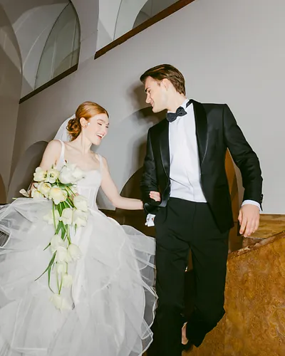 Bride in a white gown holding flowers and groom in black tuxedo walking down stairs together.