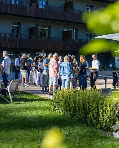 Group of people socializing outdoors near a building on a sunny day with greenery in foreground.