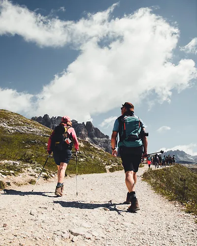 Two hikers walking on a rocky mountain trail with backpacks under a partly cloudy sky.