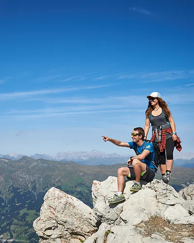 Hikers on rocky mountain peak with vast mountain range and blue sky in the background.