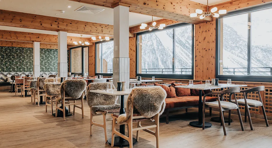 Cozy restaurant interior with wooden walls, sheepskin-covered chairs, and large windows showing snowy mountains.