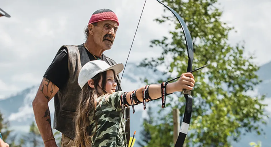 Man teaching young girl to shoot a bow and arrow outdoors with mountains and trees in the background.