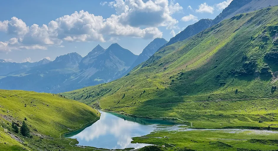 A serene mountain lake reflecting fluffy clouds with green hills and rocky peaks in the background.