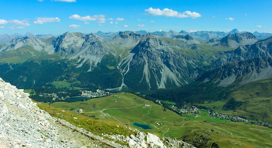 Wide mountain range with rocky peaks, green valleys, small lakes, and a clear blue sky with clouds.
