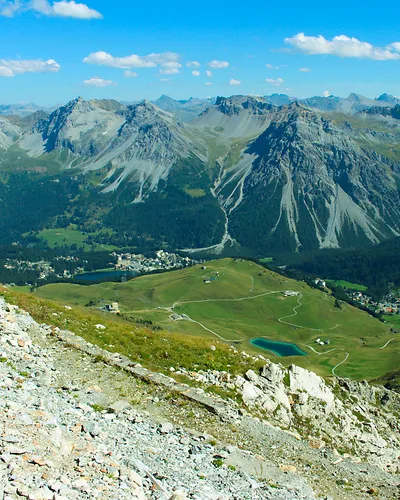 Mountain landscape with rocky foreground, green valley, small lake, and distant peaks under blue sky.
