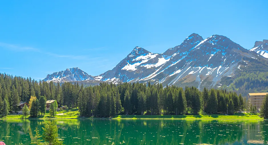 Clear lake reflecting pine trees and snowy mountain peaks under a bright blue sky.