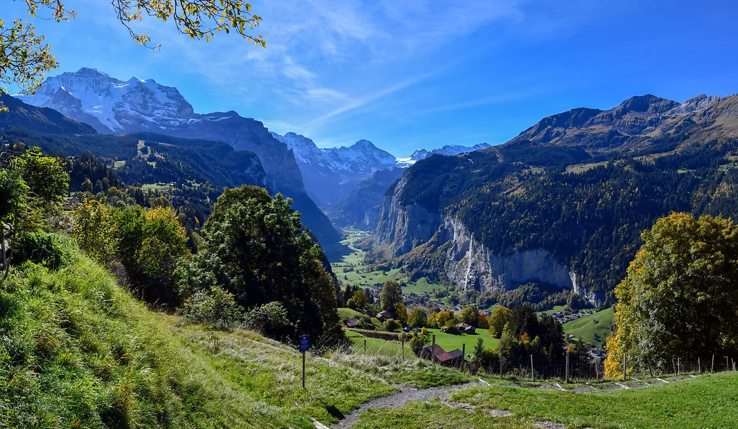 Green valley with trees, small houses, and towering snow-capped mountains under a clear blue sky.