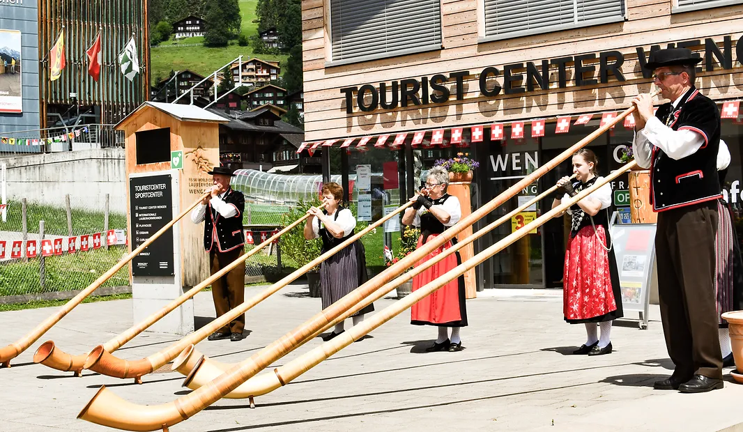Five people in traditional Swiss clothes playing long alphorns outside a tourist center.