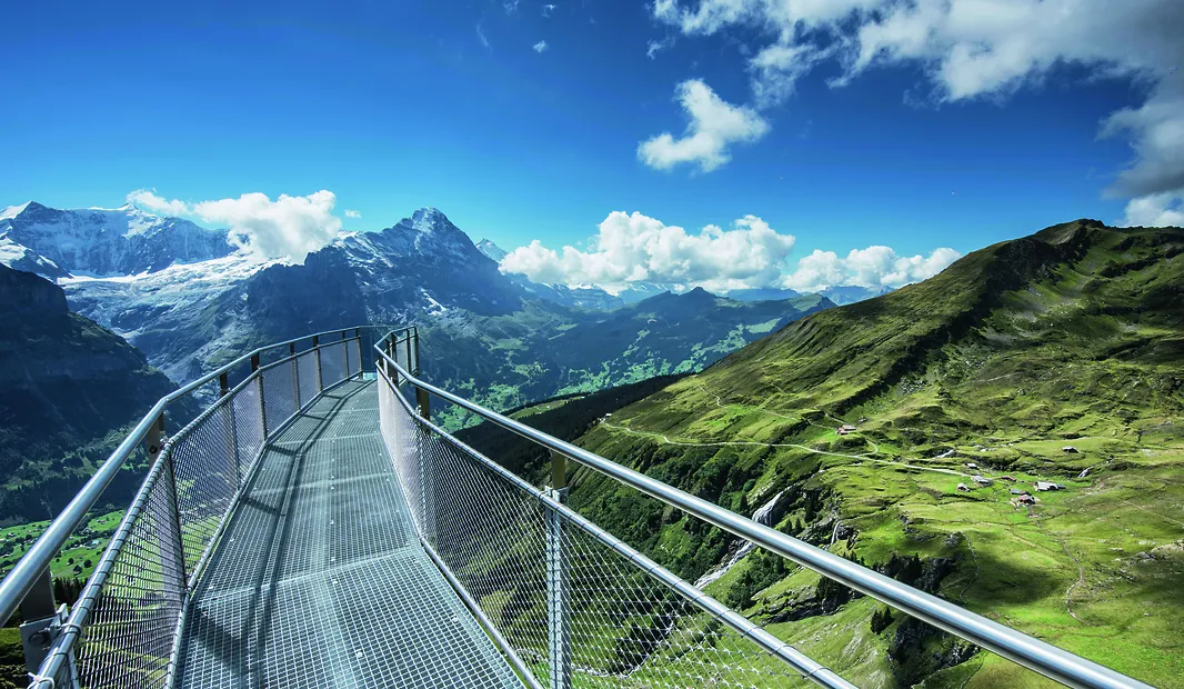Metal cliff walkway overlooking green valleys and snow-capped mountain peaks under a blue sky.