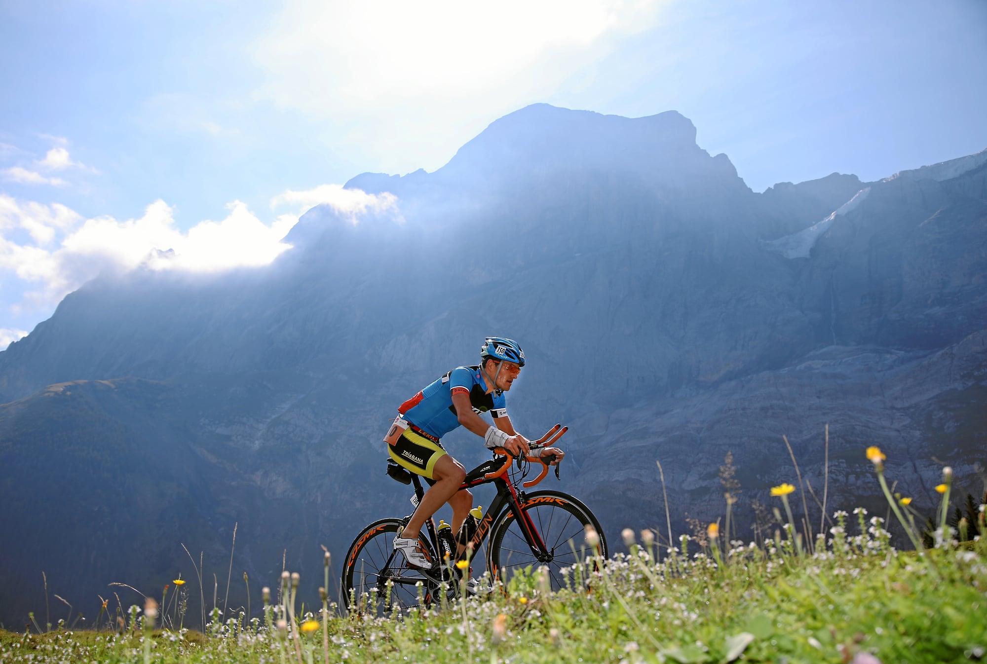 Inferno Triathlon road cyclist climbing toward Mürren with summer alpine panorama behind. Photo: © Mürren Tourismus (placeholder credit, confirm before publish).
