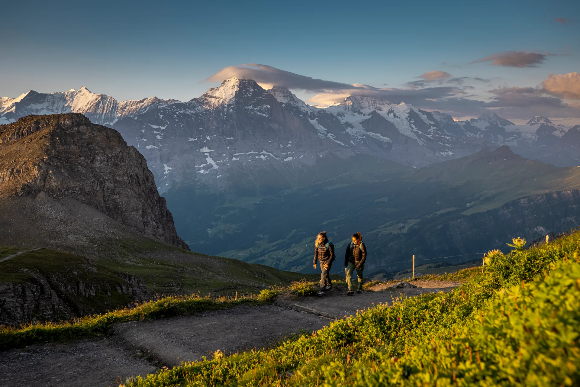 Zwei Wandernde auf einem hohen Alpenweg oberhalb von Wengen, mit den schneebedeckten Berner Alpen und einem tiefen Tal darunter