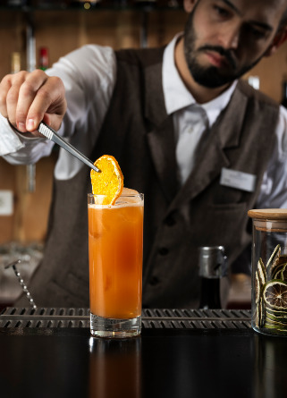 Bartender in a waistcoat garnishing a tall orange cocktail with a dried orange slice at the bar