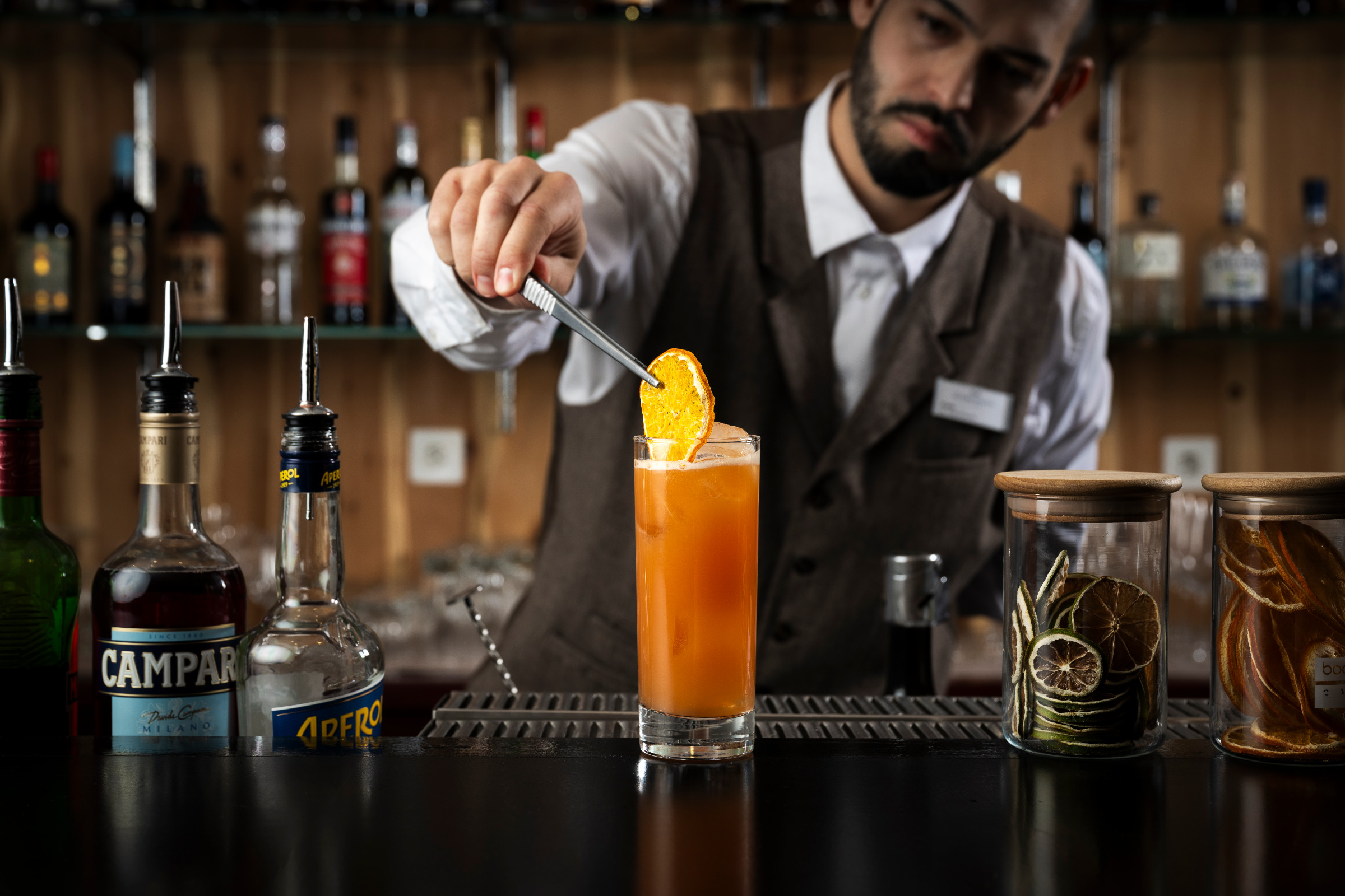 Barman en gilet garnissant au bar un grand cocktail orange d'une tranche d'orange séchée