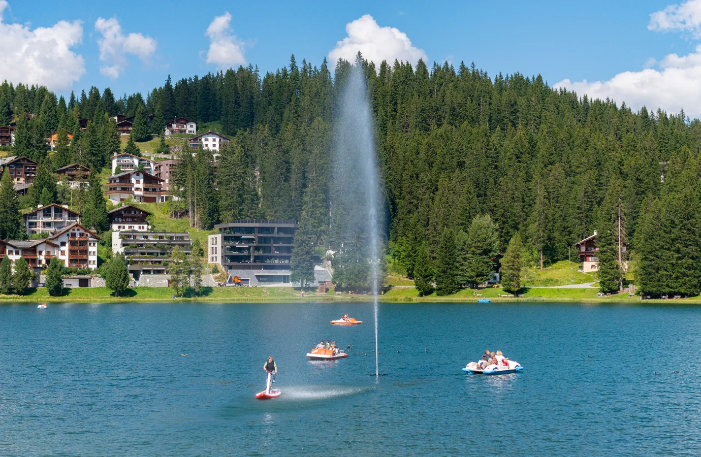 The Obersee lake in Arosa with its water fountain and the village chalets behind it on a summer day