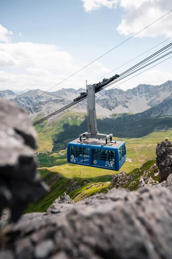 A blue Arosa Lenzerheide gondola cabin crossing above a green alpine valley in summer