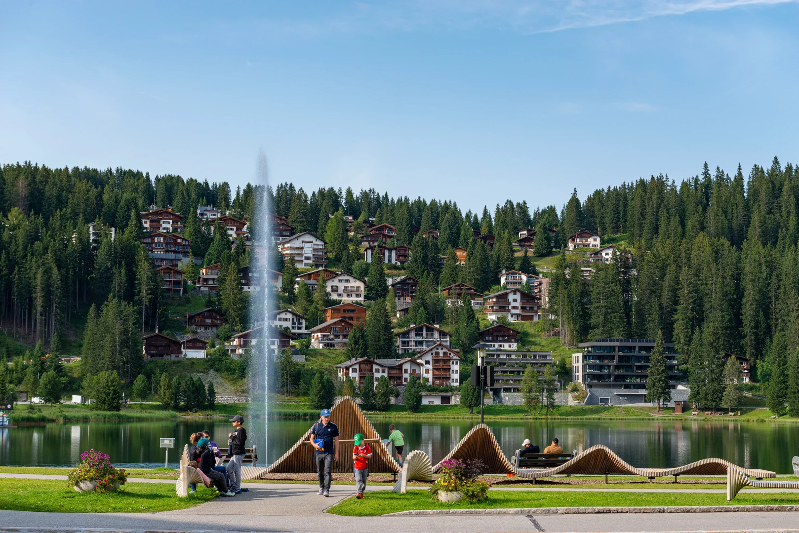 People walking the Obersee promenade in Arosa in summer with the lake fountain spraying behind them