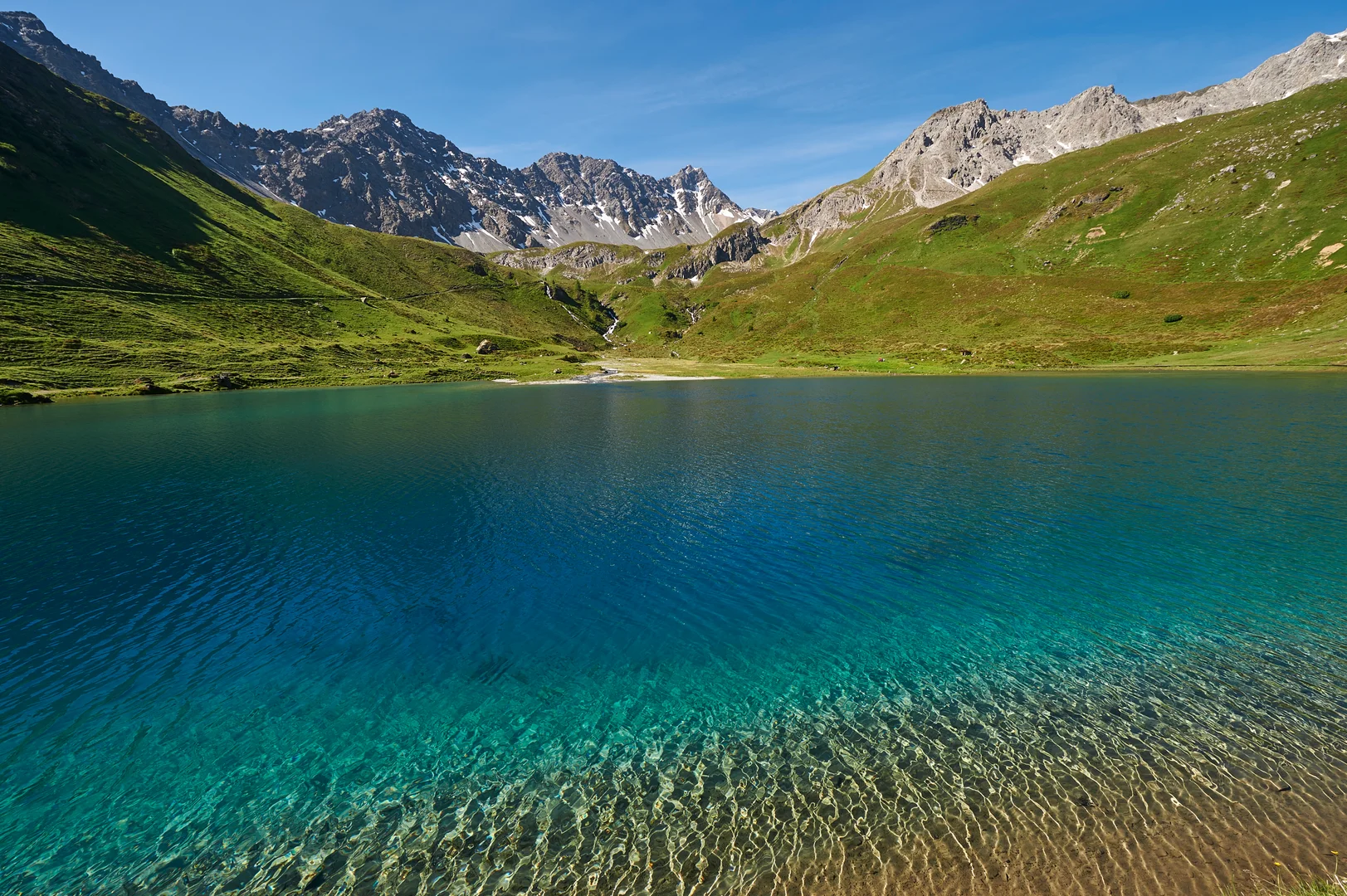 The clear turquoise water of the Schwellisee above Arosa with grassy slopes and rocky peaks behind it