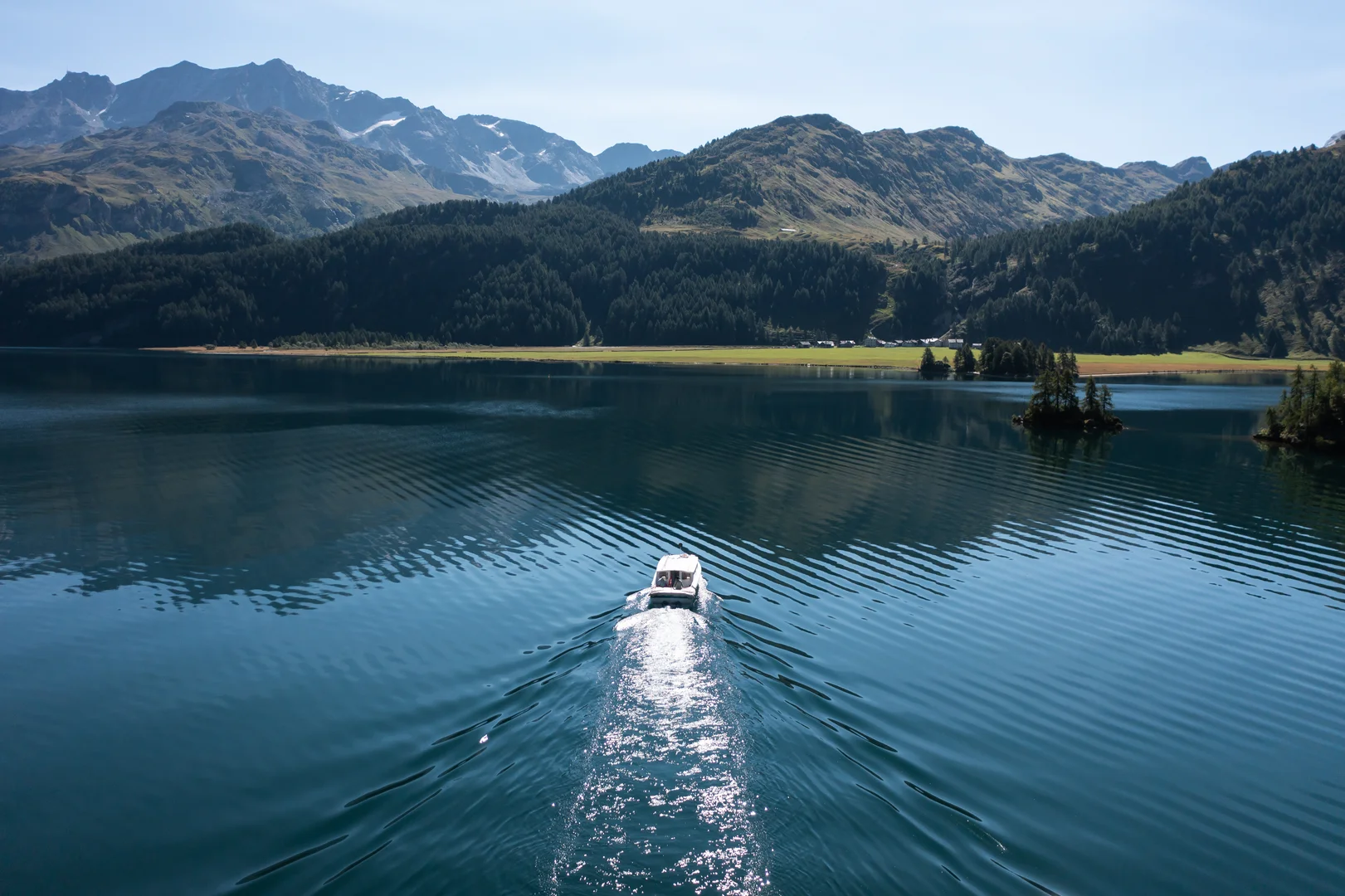 A small white passenger boat crossing Lake Sils seen from above, forested slopes and part-snowy peaks behind it