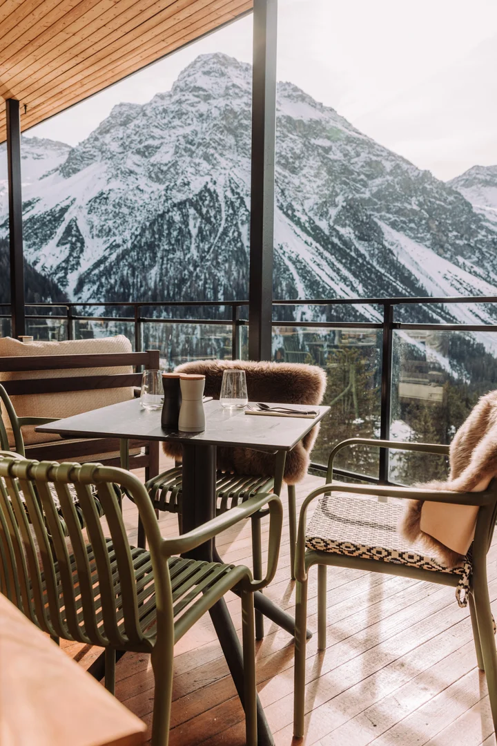 Terrasse extérieure de l’Altein, Arosa, avec une table dressée, des plaids en fausse fourrure sur des chaises à lattes et les sommets enneigés d’Arosa au-delà du garde-corps vitré