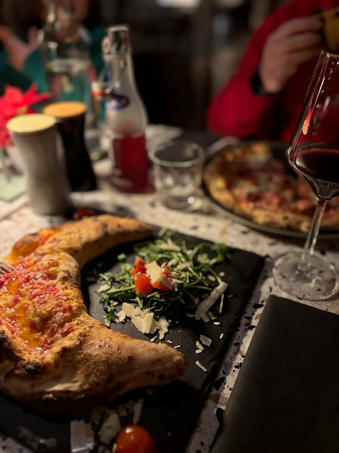 Une pizza au feu de bois sur une planche en bois, posée sur une table de restaurant éclairée à la bougie à Arosa, avec un verre de vin rouge à côté