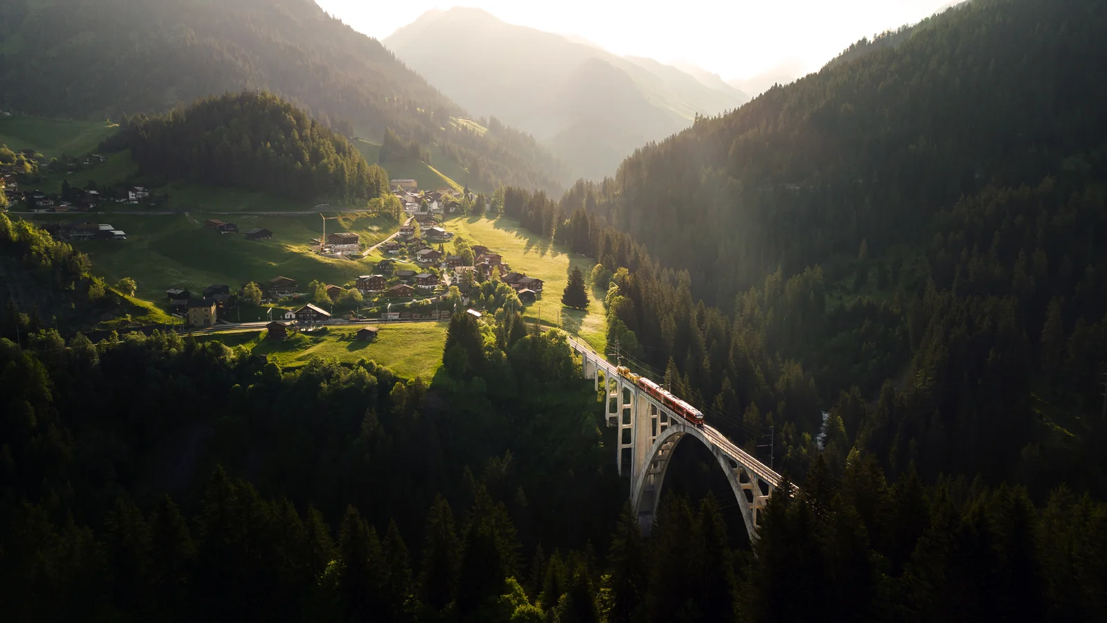 A red Rhaetian Railway train crossing a tall curved stone viaduct above a wooded valley in summer