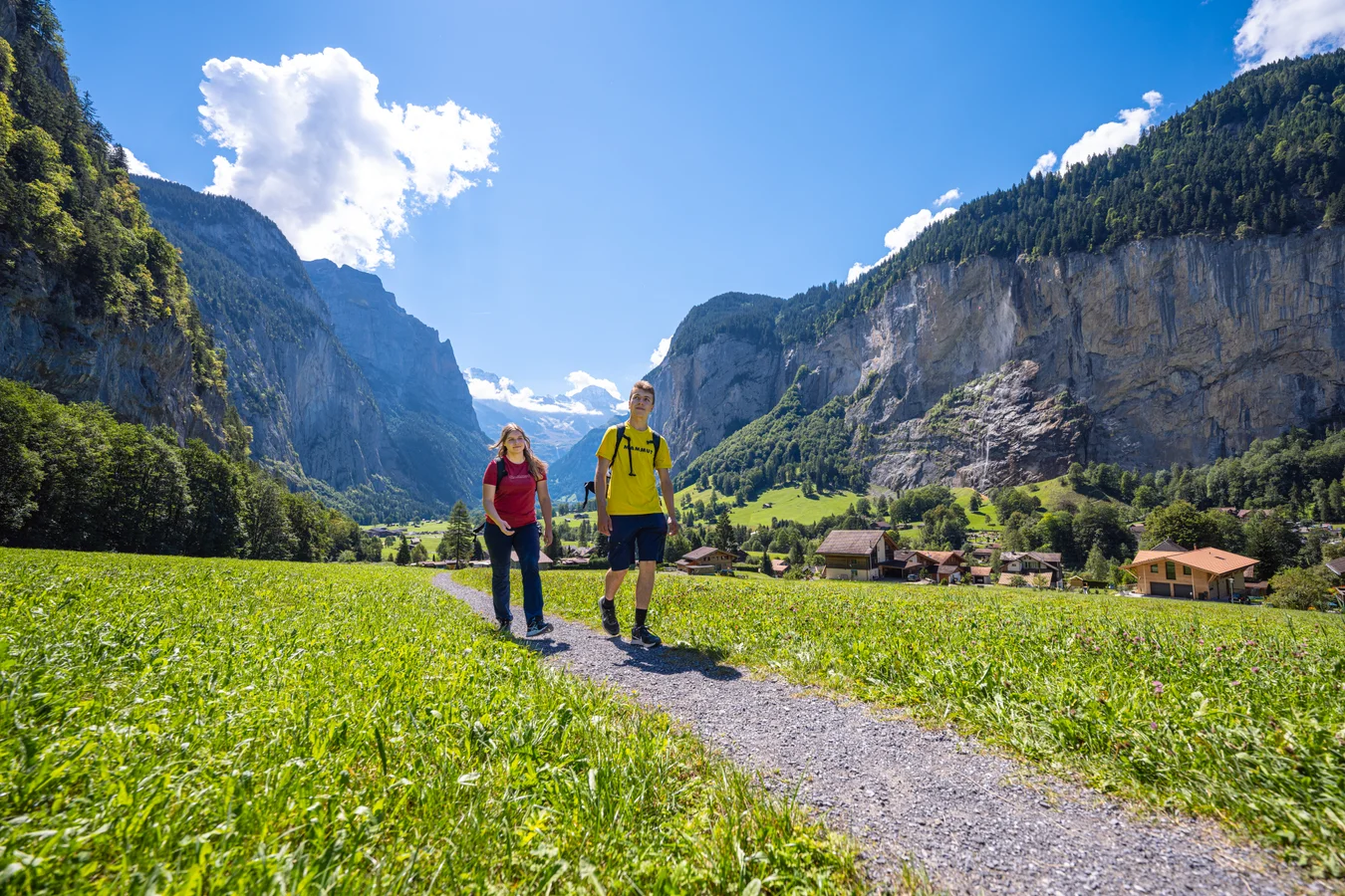 Zwei Wanderer auf einem Kiesweg im Talboden des Lauterbrunnentals, beidseits steile Felswände und ein Dorf an deren Fuss