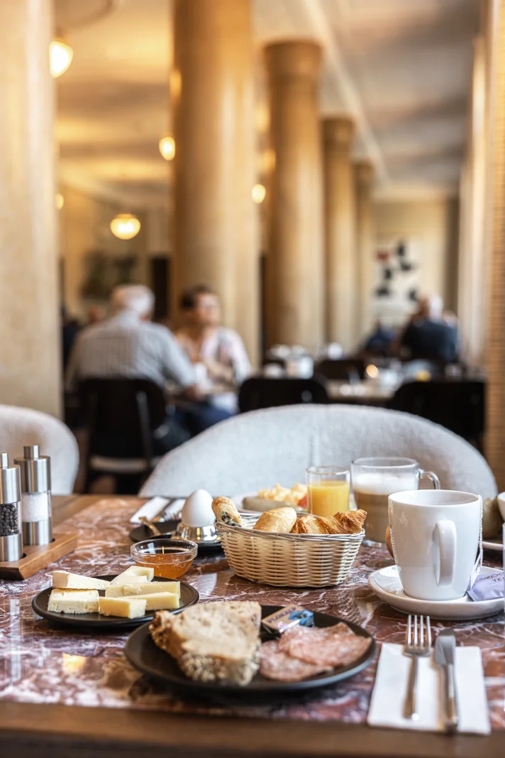 Table with breakfast items including cheese, bread, cold cuts, croissants, boiled egg, coffee, and juice in a restaurant.