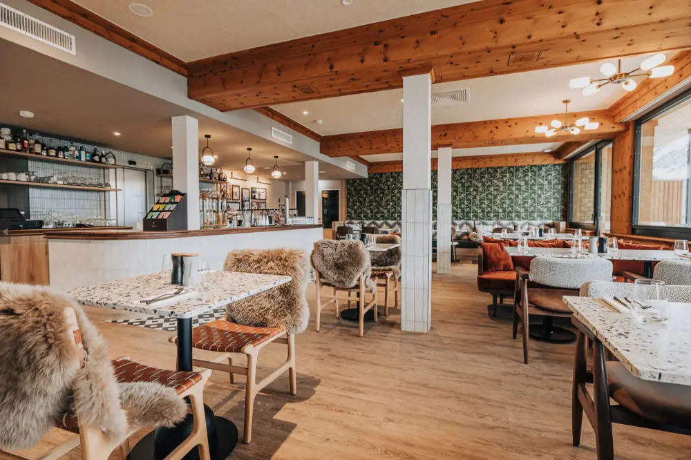Modern restaurant interior with wooden beams, fur-covered chairs, patterned walls, and set tables.
