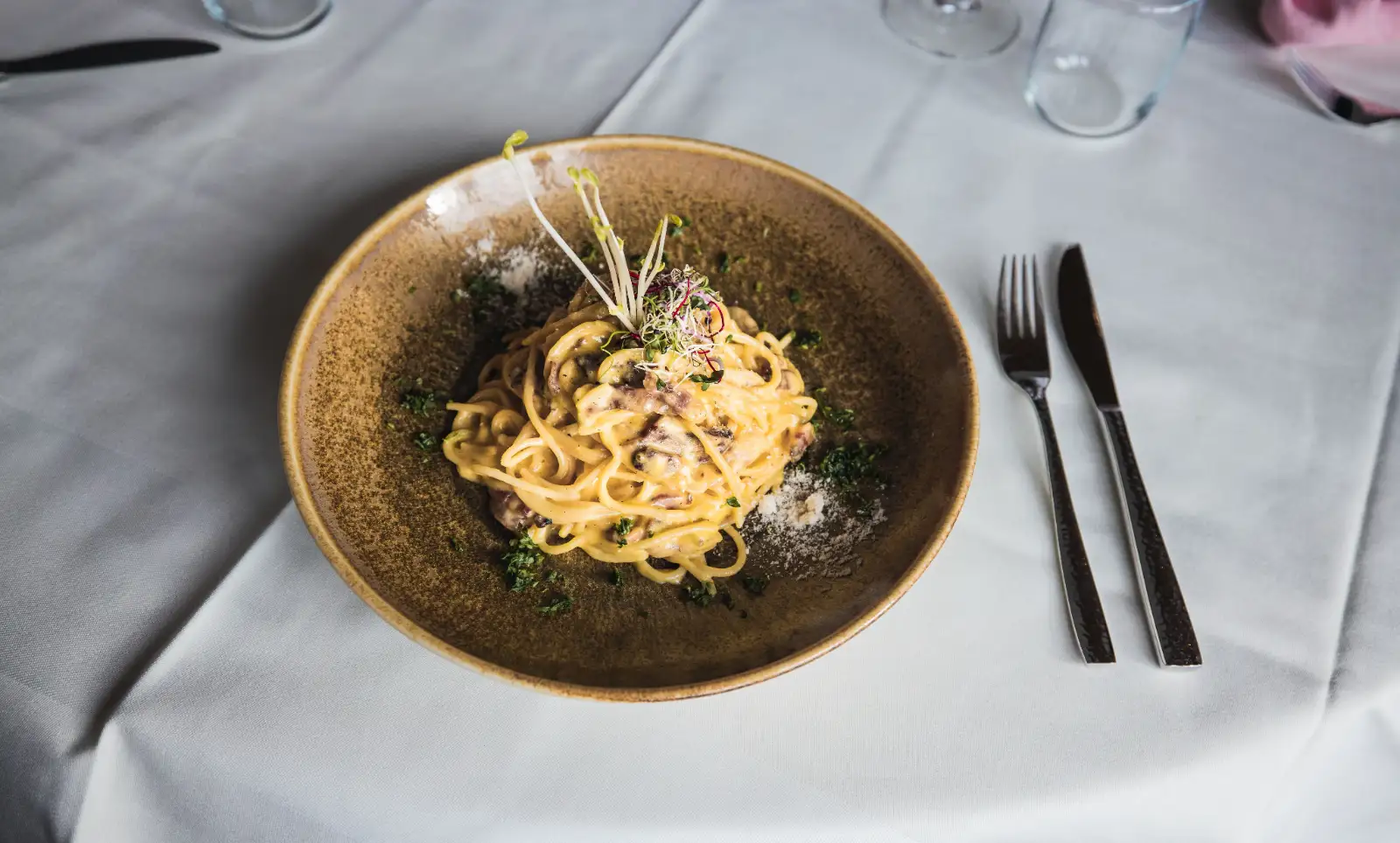 Plate of creamy pasta with mushrooms garnished with herbs on a white tablecloth, with knife and fork.