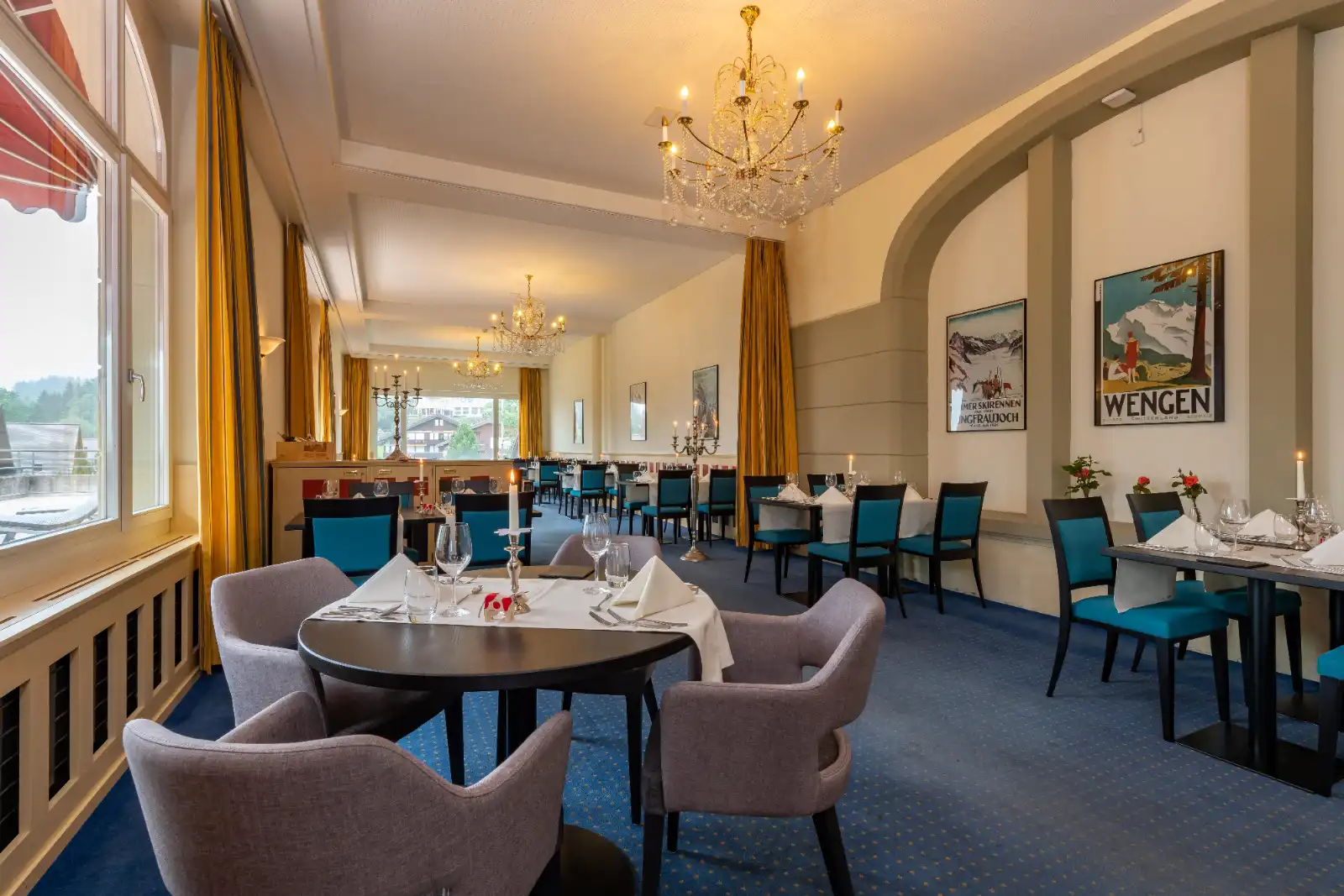 Elegant dining room with chandeliers, blue and gray chairs, white tablecloths, and vintage Wengen posters.