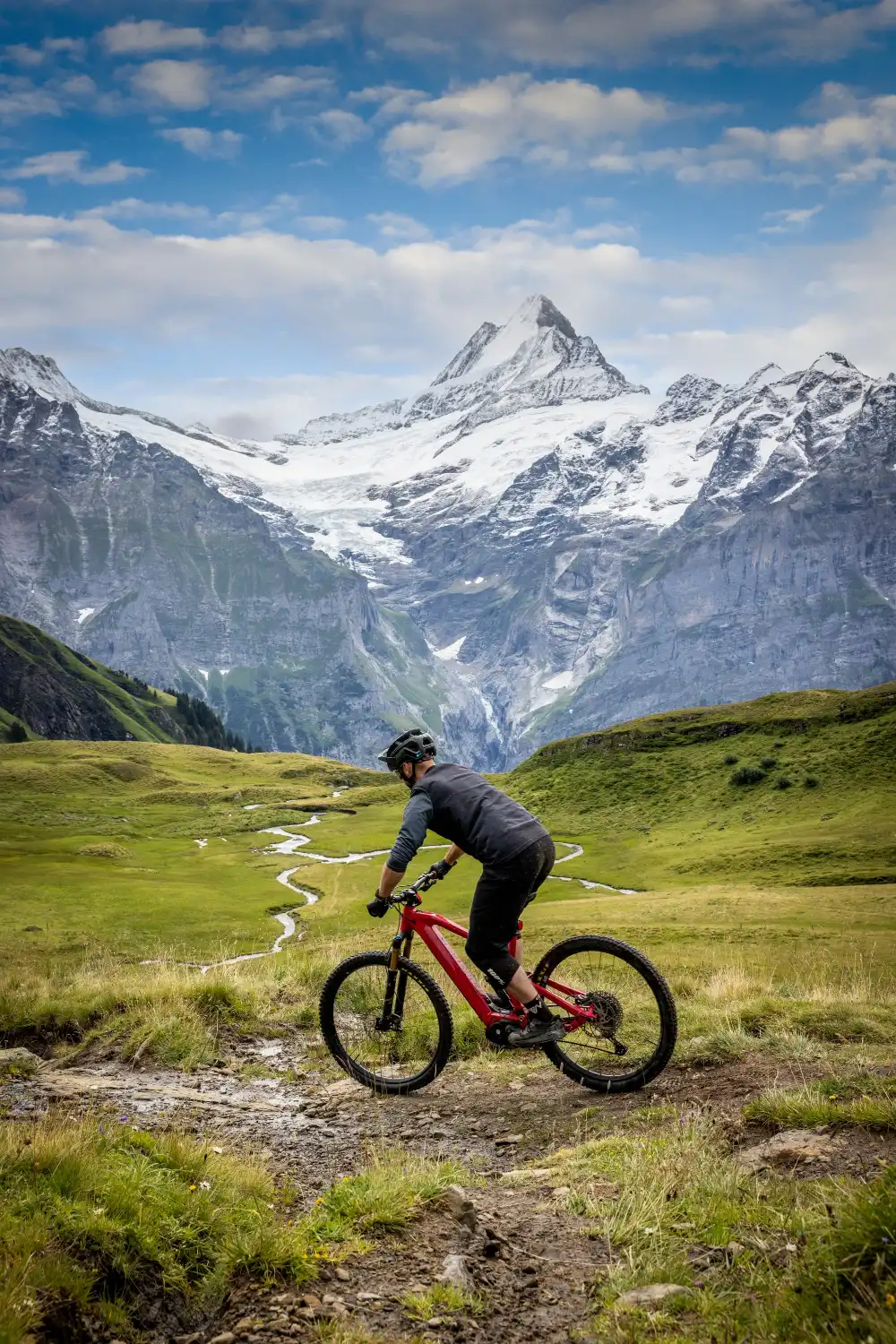 Mountain biker on a red bike riding a dirt trail through alpine meadow with the Wetterhorn behind near Grindelwald. Photo: © Grindelwald Tourismus (placeholder credit, confirm before publish).