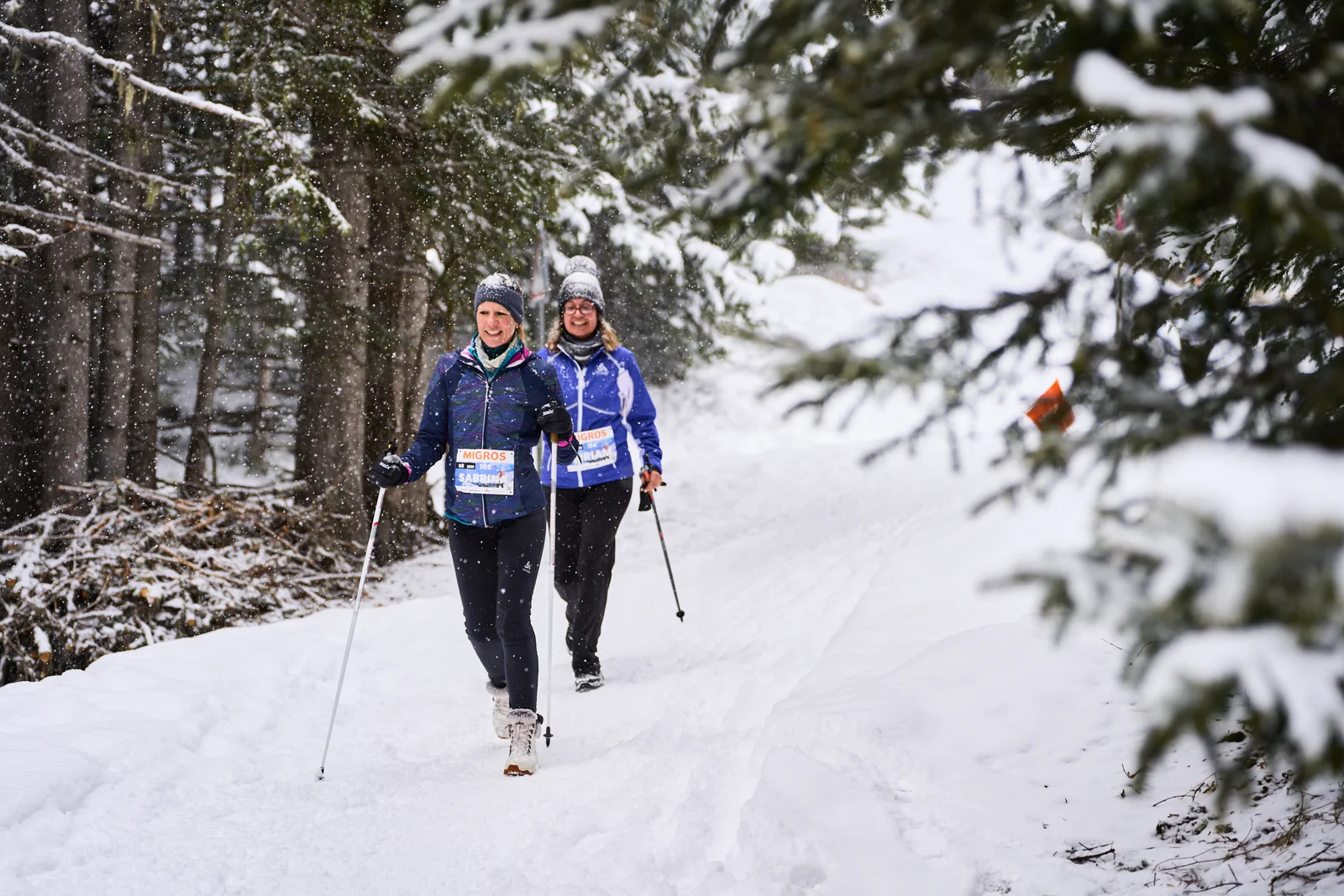Two walkers with poles on a snowy forest path near Arosa in falling snow, an orange flag at the edge