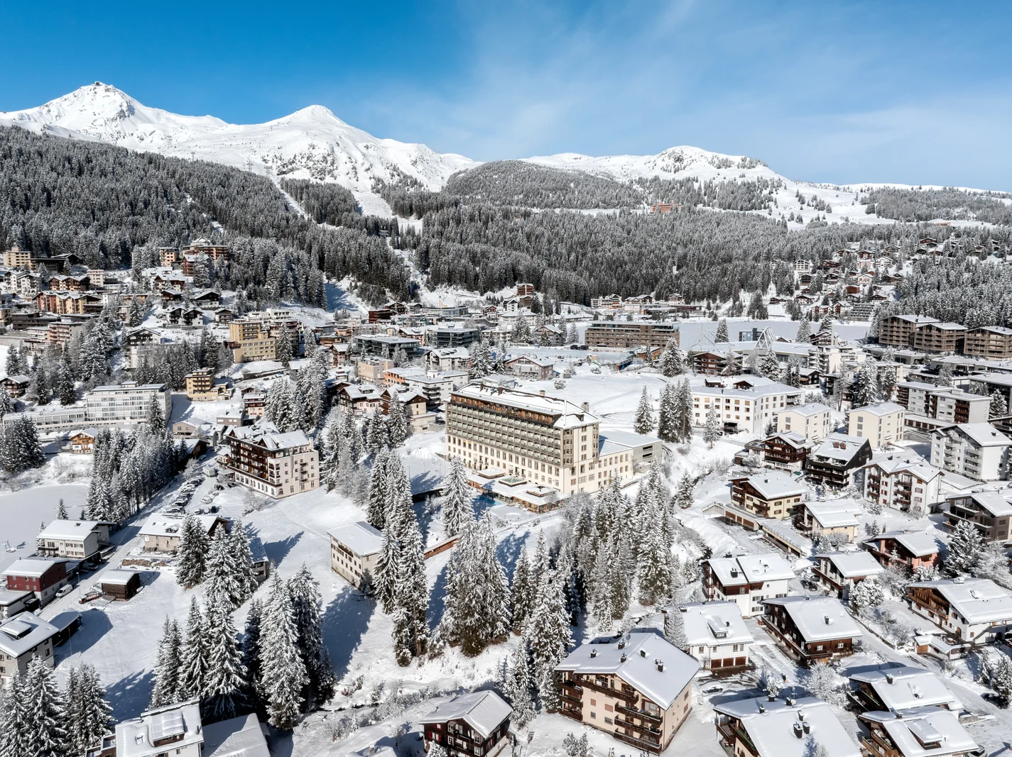 Tetti innevati e abeti di Arosa visti dall'alto, con un lago ghiacciato in alto a destra