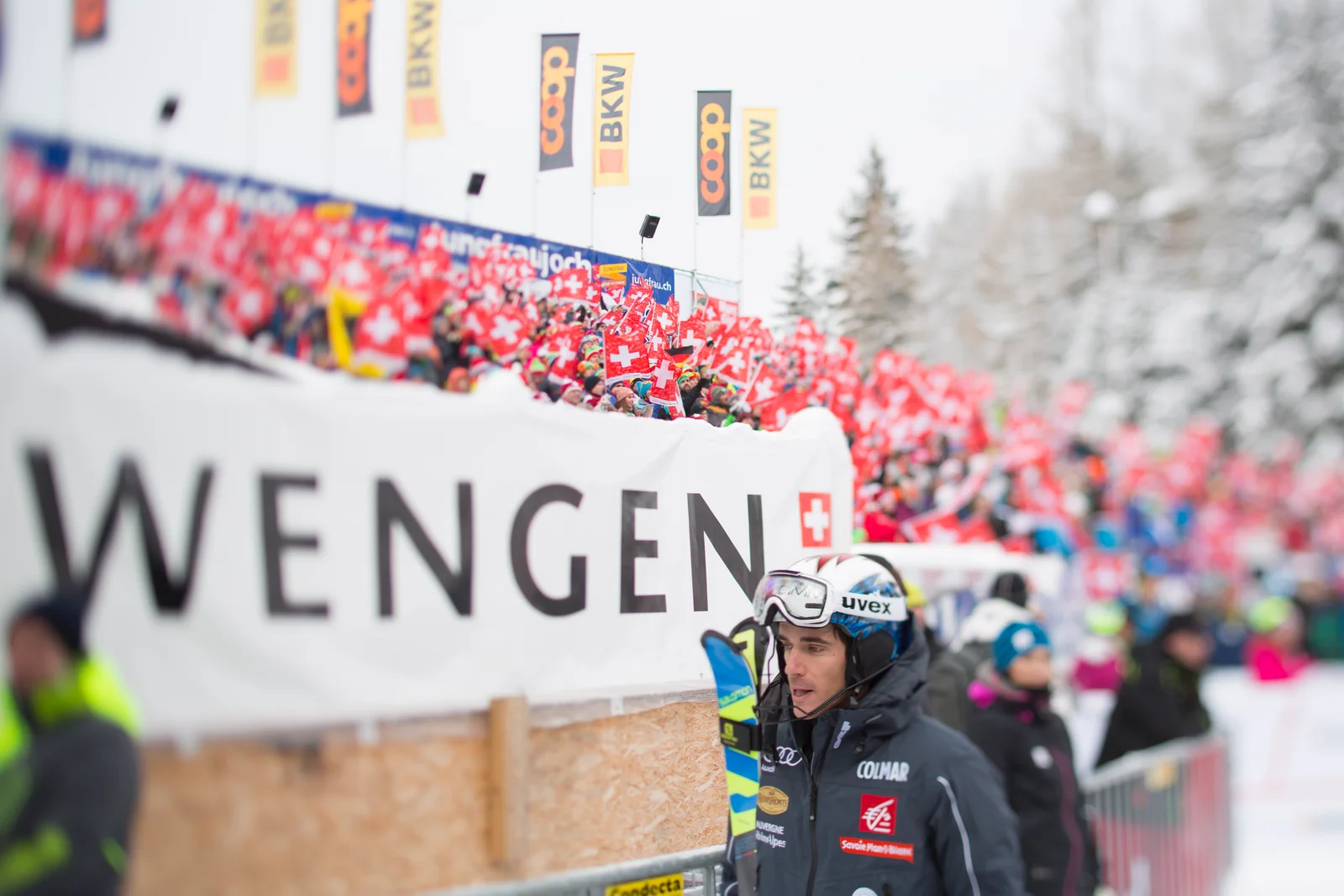 Racer in the Lauberhorn finish area below a WENGEN banner, with a stand of fans waving Swiss flags behind