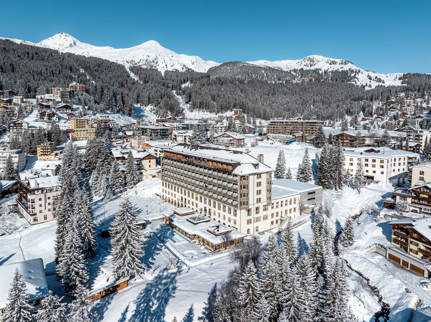 Hotel Altein and neighbouring buildings in snow, seen from above with forested slopes of Arosa beyond