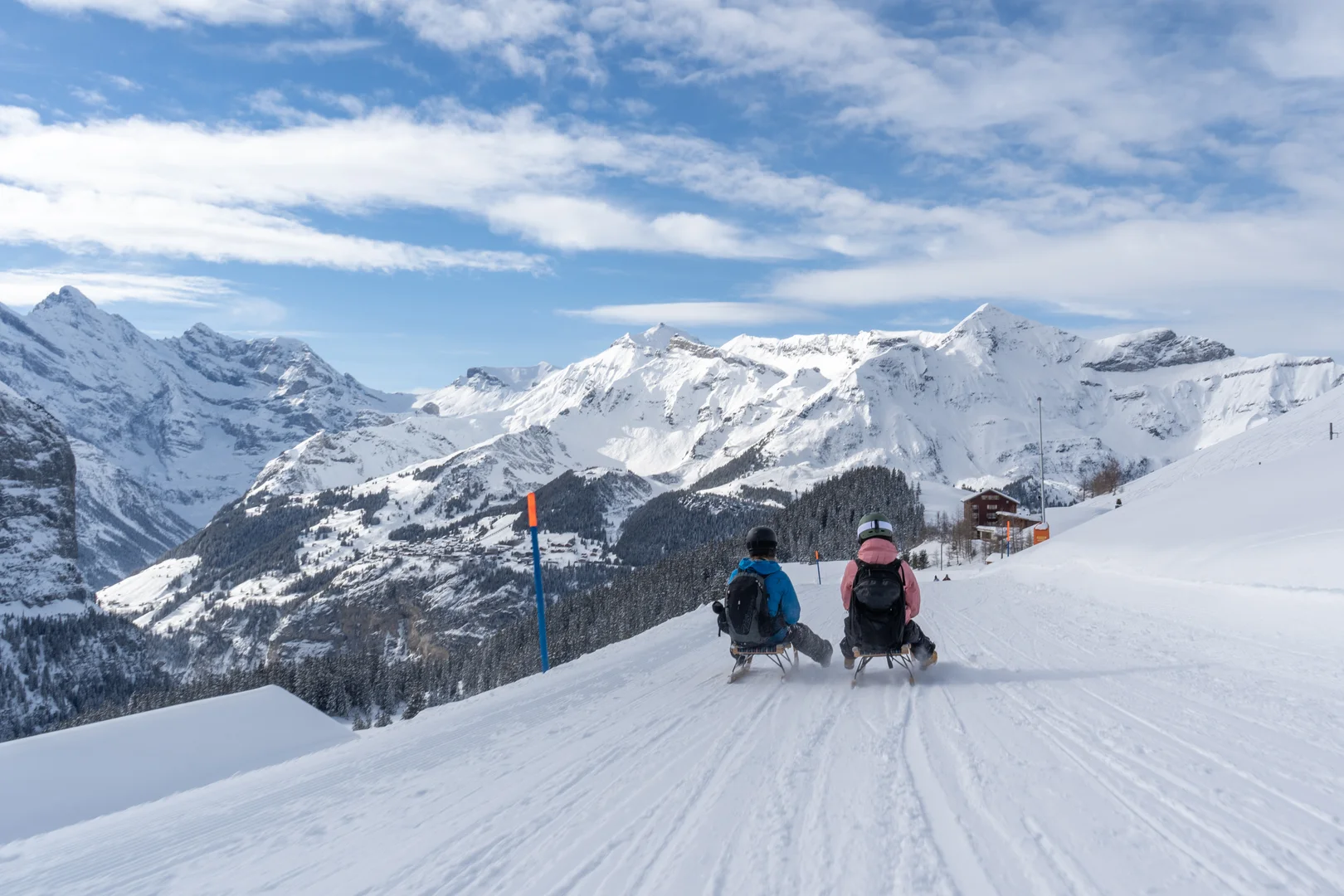Due slittinisti su una pista da slittino battuta, con cime innevate e una baita sullo sfondo