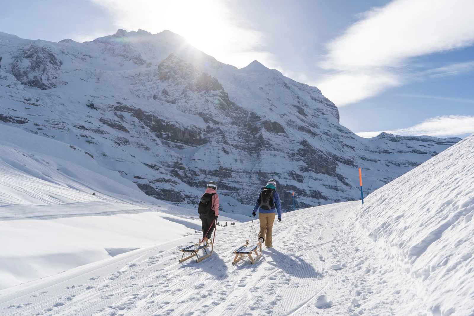 Due persone trainano slittini di legno su un sentiero innevato, sotto una ripida parete rocciosa
