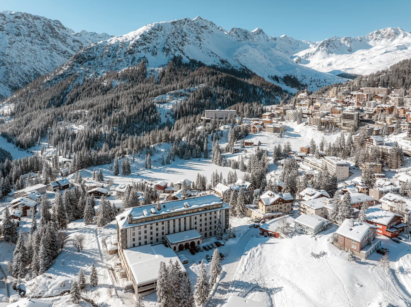 Aerial view of Hotel Altein among snowy firs in Arosa, with forested slopes and white peaks behind