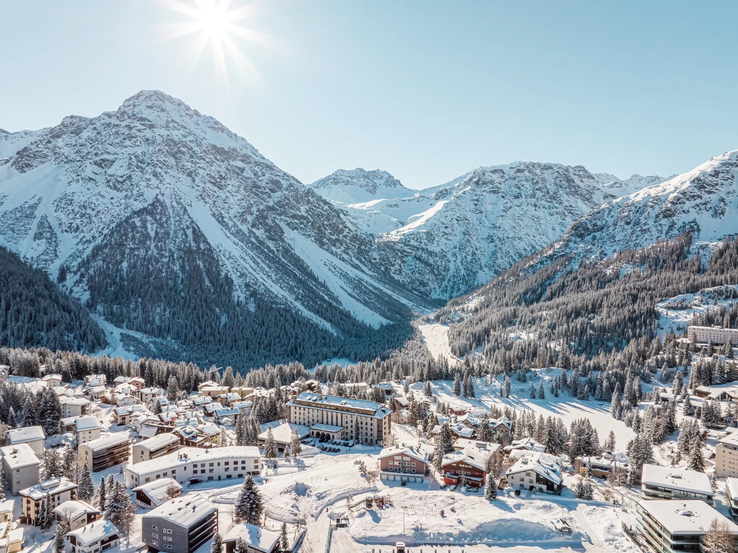 Veduta aerea di Arosa innevata al sole, con pendii boscosi e cime innevate sopra il paese