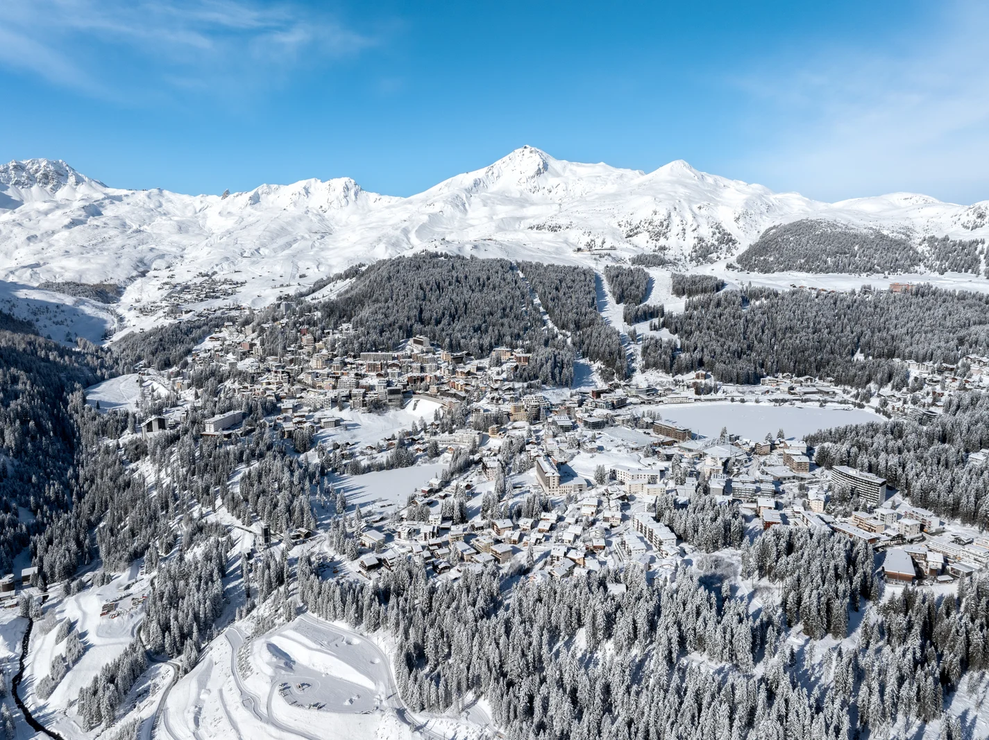 Winter aerial of Arosa with a frozen lake in the centre, snow-covered forest and ski slopes around it