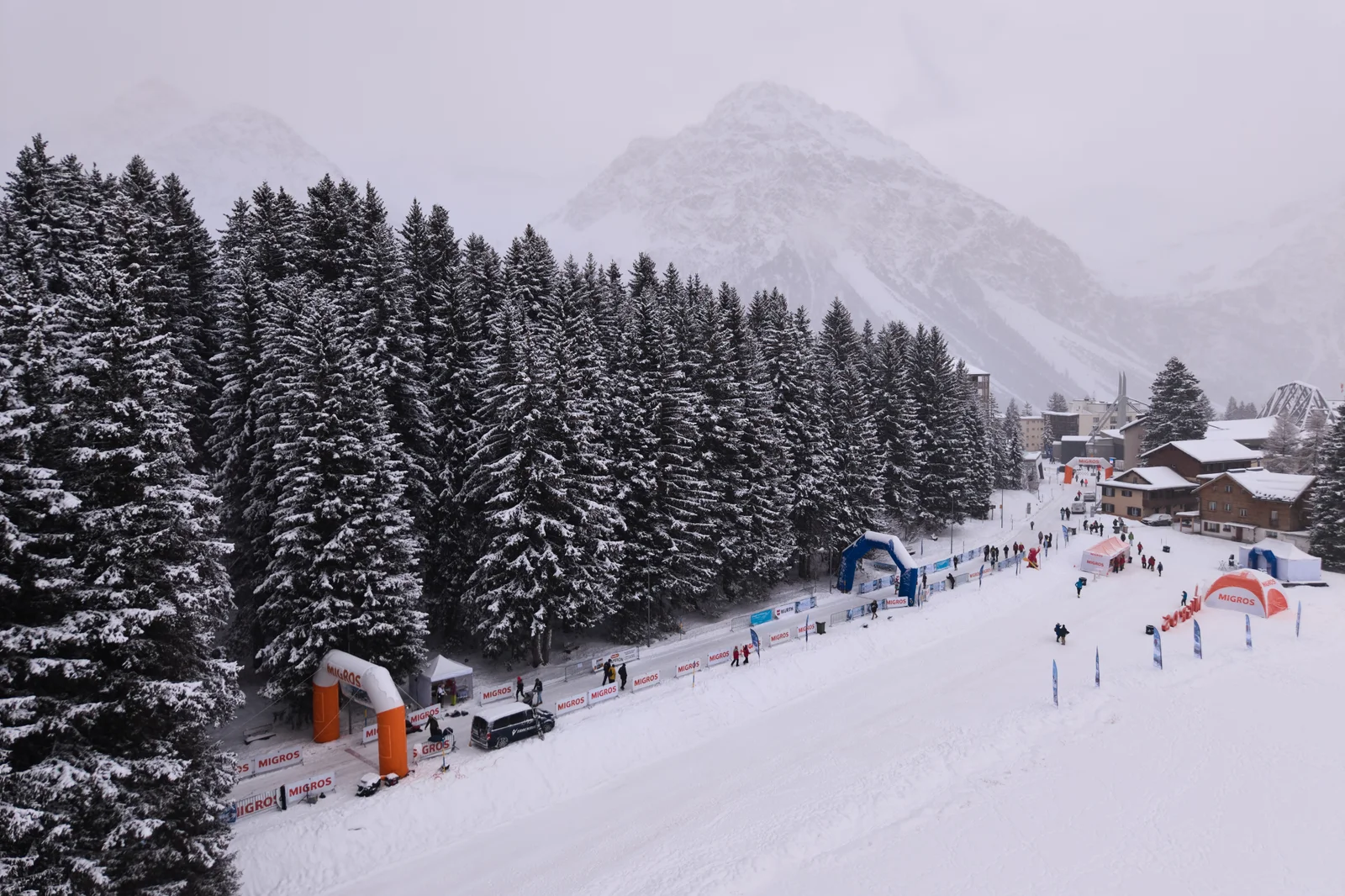Veduta aerea di un'ampia pista battuta con archi di un evento accanto a un bosco innevato, chalet e cime dietro