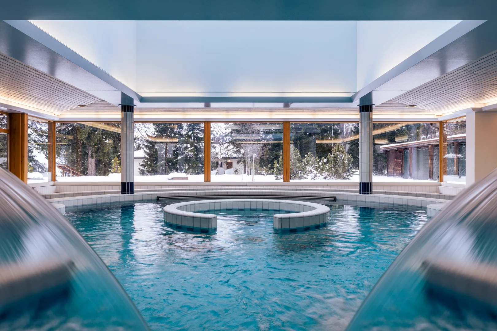 Piscina coperta dell'Altein, Arosa, con seduta curva piastrellata e vetrate panoramiche sul paesaggio innevato con abeti