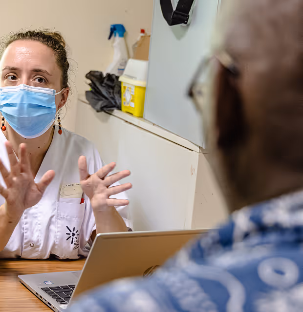 A woman wearing a mask gestures while speaking to a person across a desk.