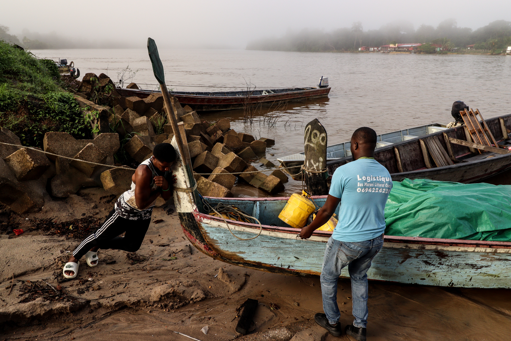 Two men work together to pull a boat ashore from a riverbank.