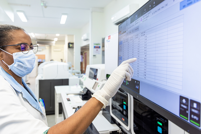 A technician wearing a mask and gloves points at data on a large screen in a lab.