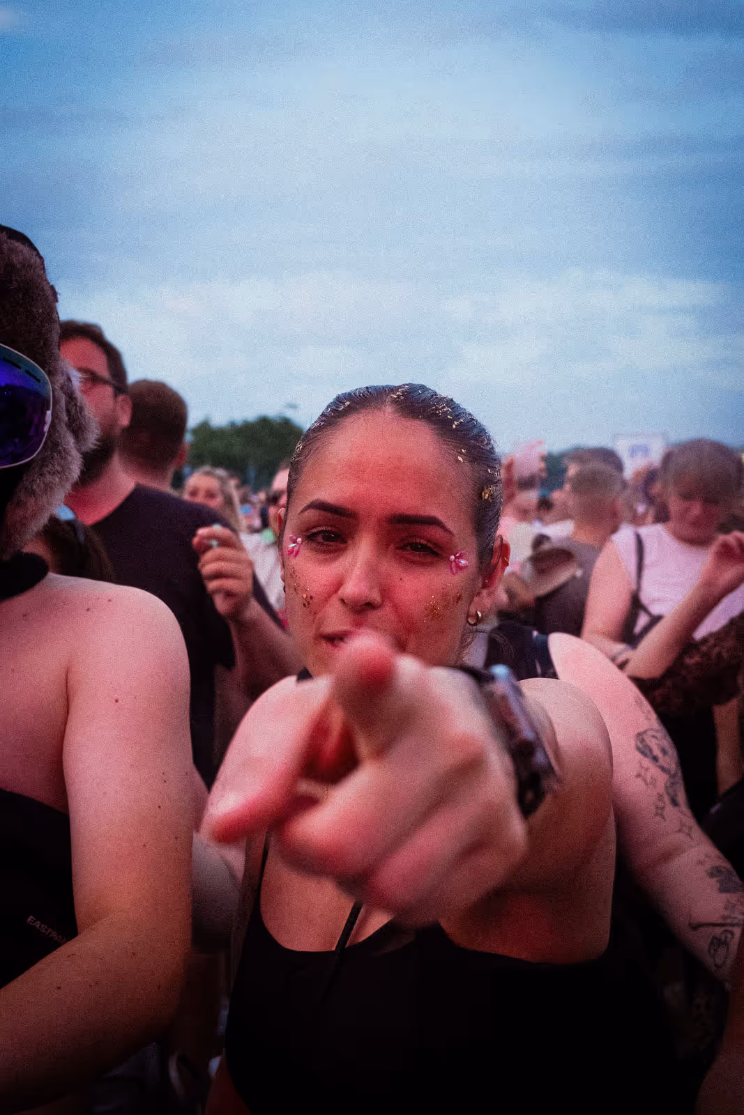 Frau mit Glitzerdekoration im Gesicht zeigt direkt in die Kamera auf einem Outdoor-Festival mit Menschen im Hintergrund.