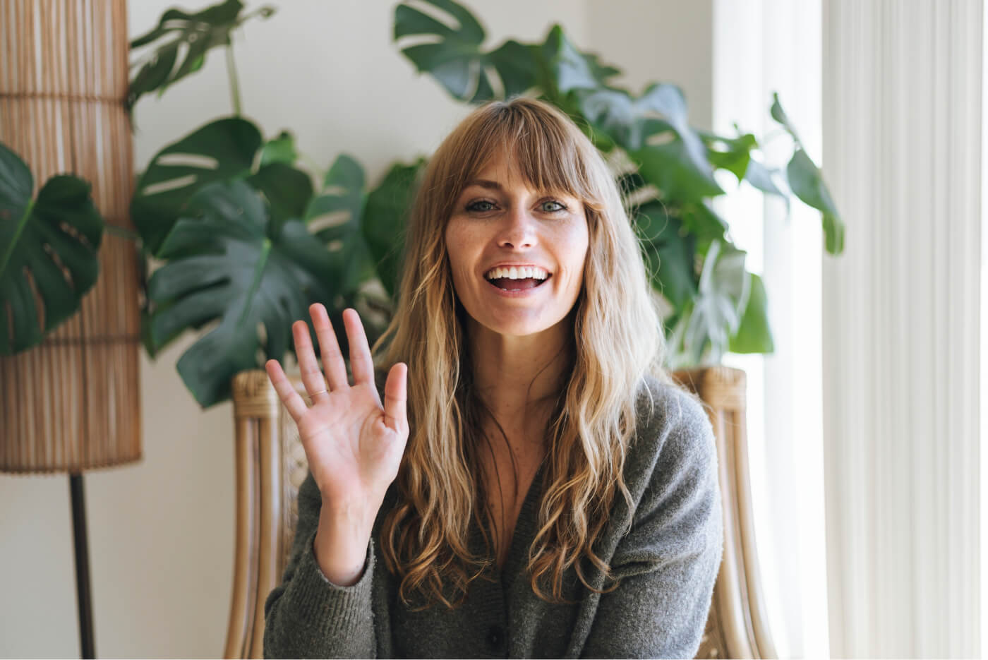 Smiling woman with long blonde hair wearing a gray sweater, waving at the camera indoors with green plants in the background.