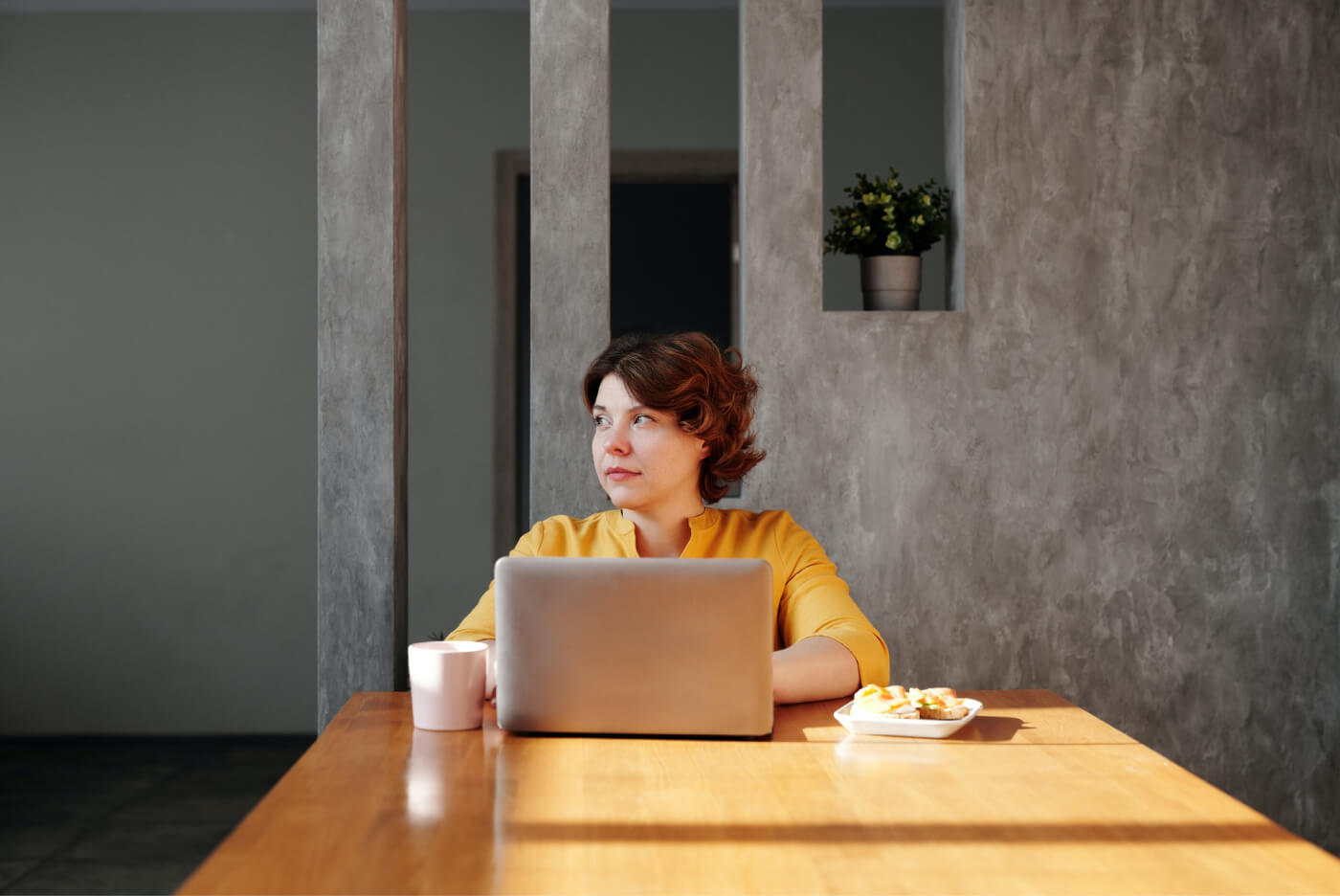 Woman in yellow shirt sitting at a wooden table with a laptop, cup, and plate of food, looking to the side.