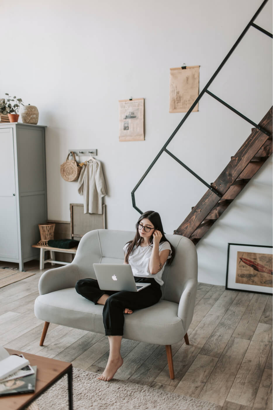 Woman with glasses sitting barefoot on a gray sofa using a laptop in a cozy modern living room with wooden stairs and light decor.