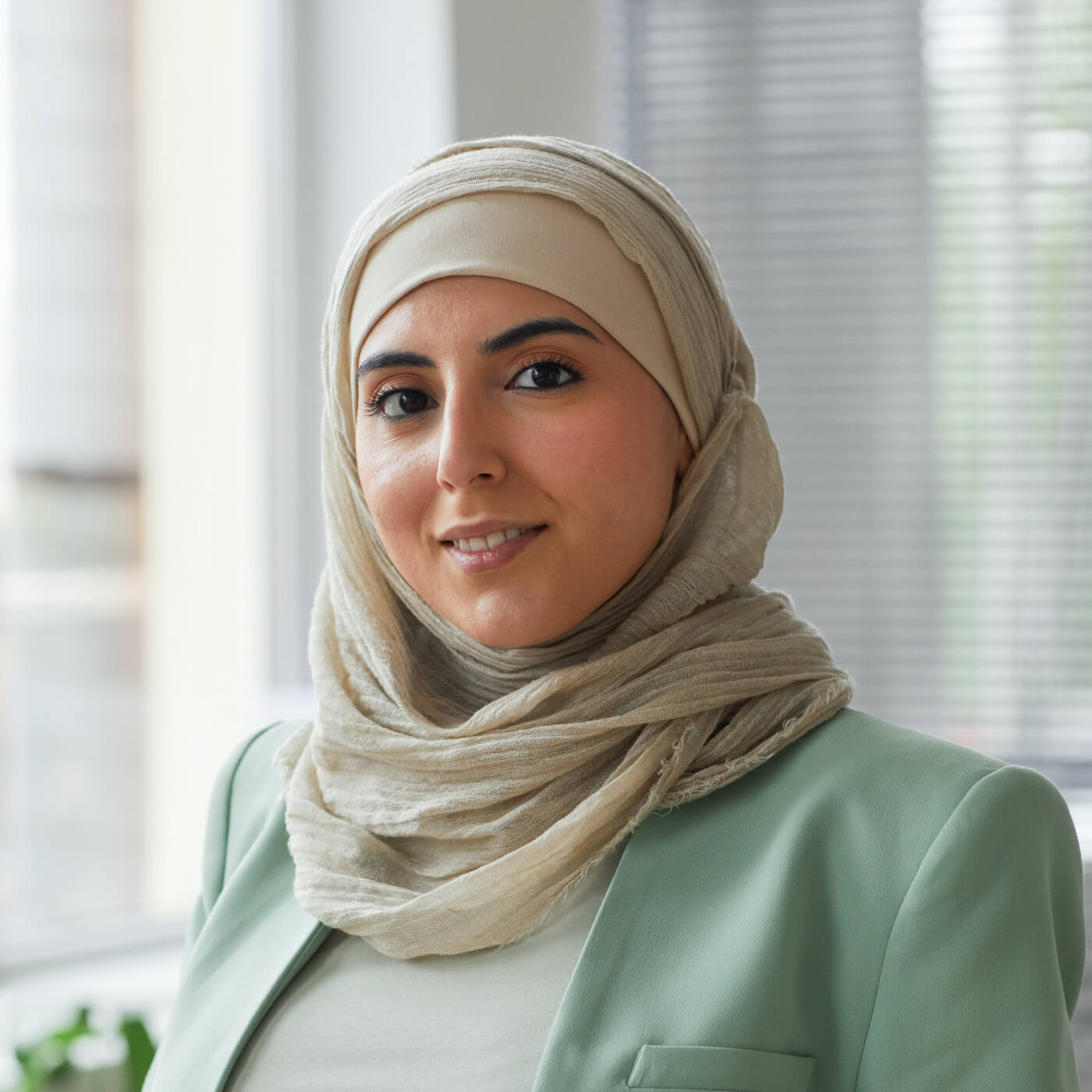 Confident woman wearing a beige hijab and light green blazer smiling indoors.